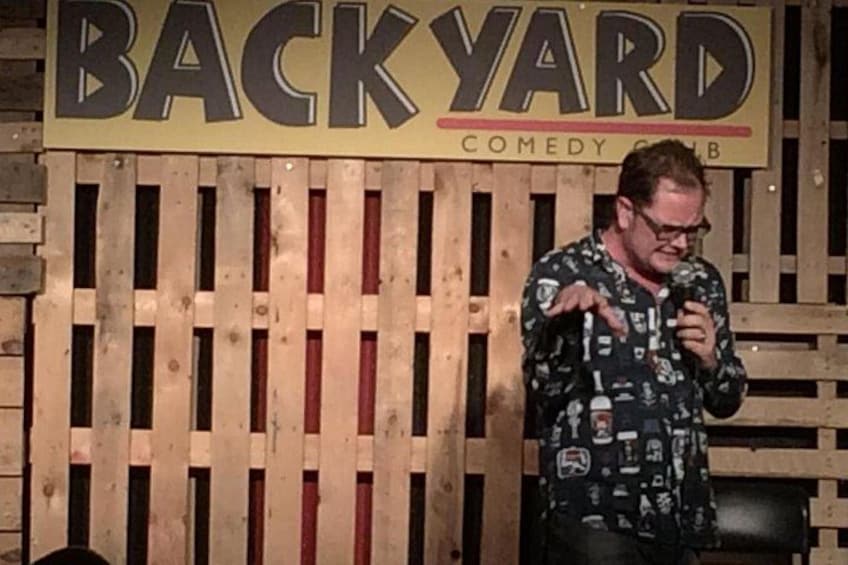 Un homme portant une chemise à motifs se tient devant une clôture en bois, sur laquelle est affiché un panneau indiquant « BACKYARD COMEDY CLUB ». 2