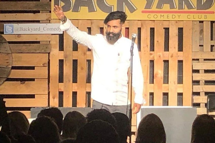 Une personne se tient sur une scène, un micro à la main, devant une clôture en bois et une pancarte sur laquelle on peut lire : « BACKYARD COMEDY CLUB ». 4