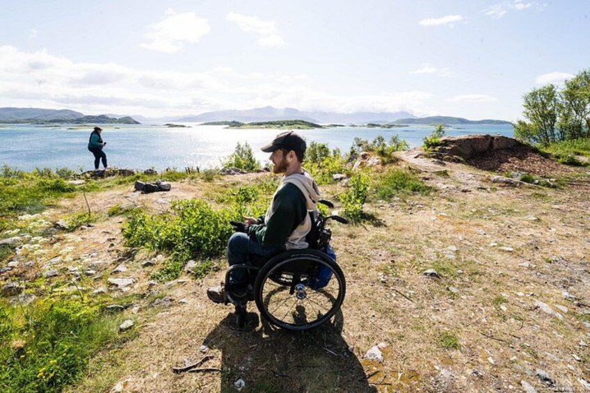 A person in a wheelchair by a lake with a person fishing in the background. 4