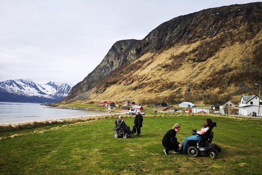 A grassy area with people in wheelchairs, a body of water, and houses in the background. 1
