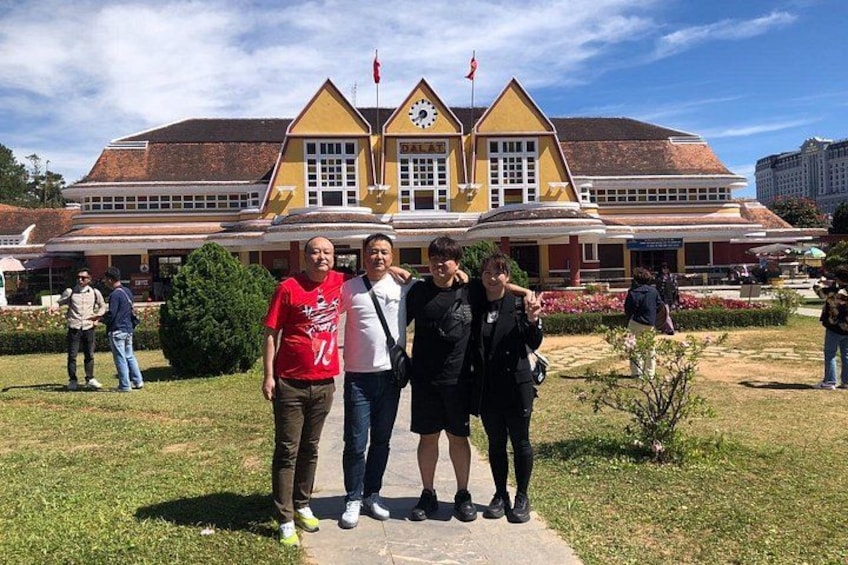 A group of people standing in front of a yellow building with a clock and flags. 4