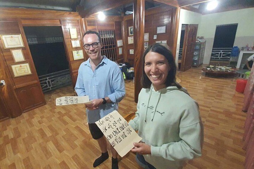 Deux personnes tenant des pancartes dans une pièce dont le sol et les murs sont en bois, avec quelques certificats accrochés aux murs et un distributeur d’eau en arrière-plan. 2