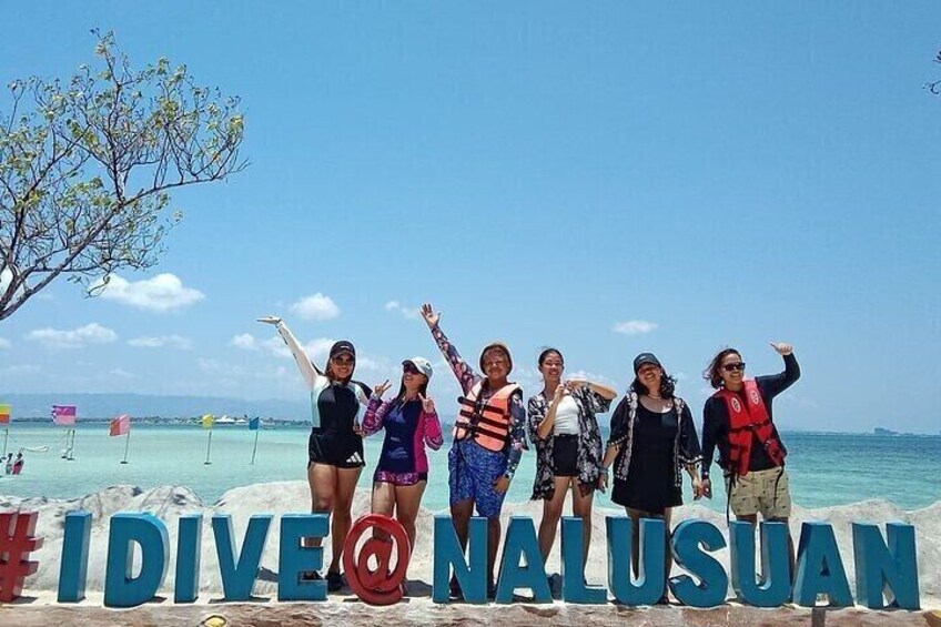 A group of people standing in front of a sign that reads "DIVINE NALUSUAN" on a beach with clear skies and calm waters. 1