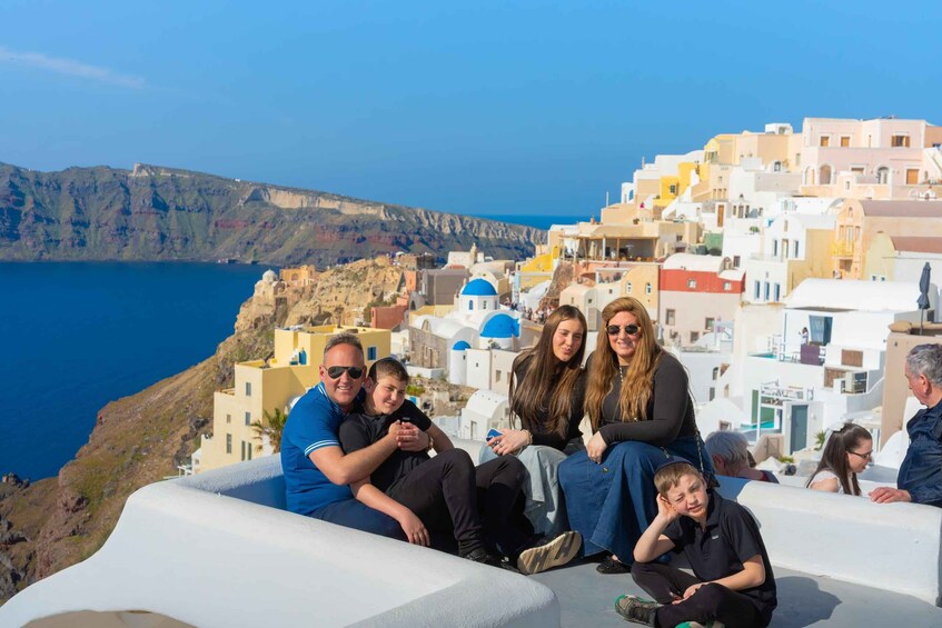 A group of people sitting on a white structure overlooking a coastal town with white buildings and blue domes, with a mountainous backdrop. 2