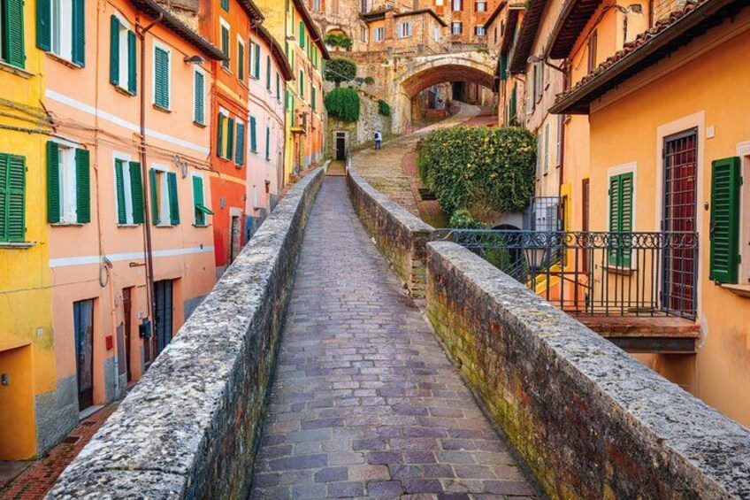 Una estrecha calle adoquinada bordeada por edificios coloridos con contraventanas verdes, que conduce a un puente de piedra. 2