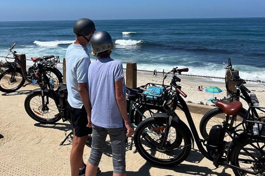 Dos personas de pie junto a una playa con bicicletas y el océano al fondo. 1