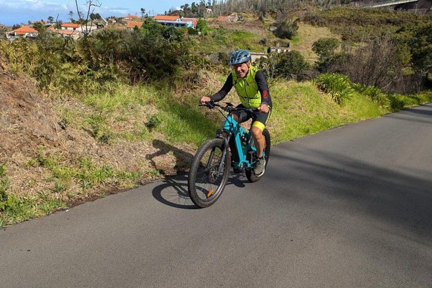 A person riding a blue bicycle on a paved road with greenery and houses in the background. 5