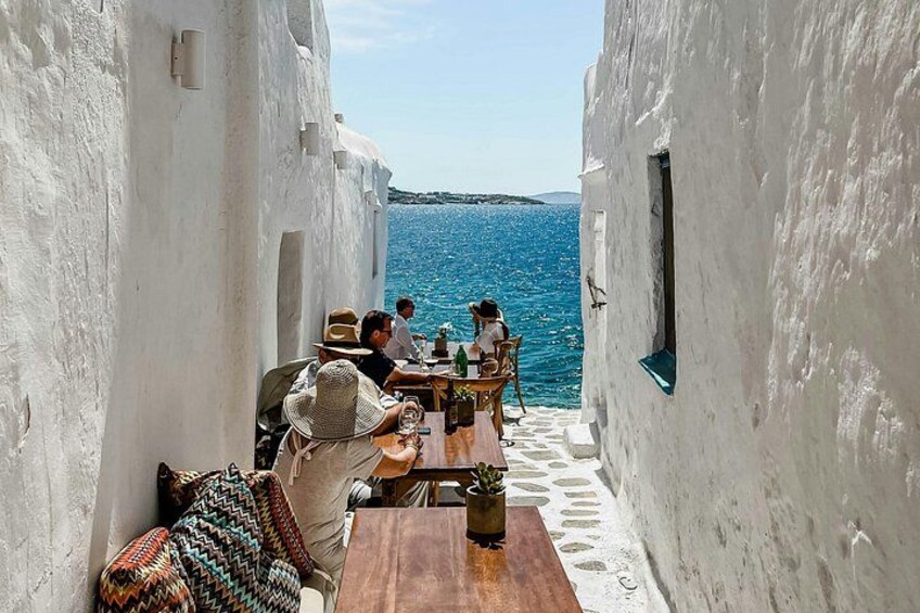 A narrow alley with white walls, a wooden table, and a view of the sea. 1