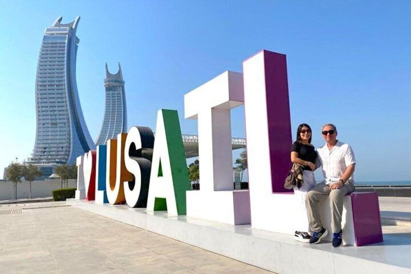 A couple sitting in front of a large 'VILUSA' sign with modern buildings in the background. 1