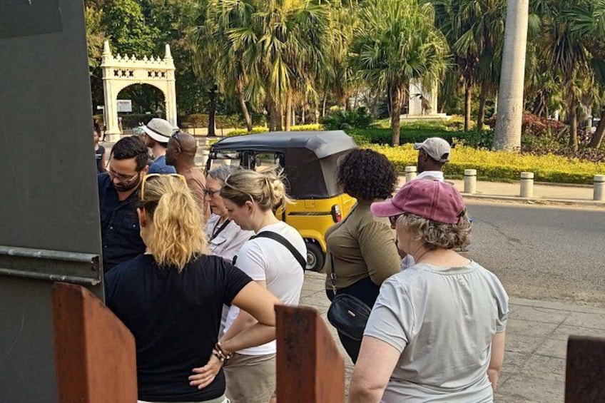 A group of people standing near a yellow auto-rickshaw, with a park and a white archway in the background. 3