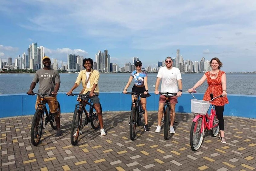 Five people on bicycles by a waterfront with a city skyline in the background. 1