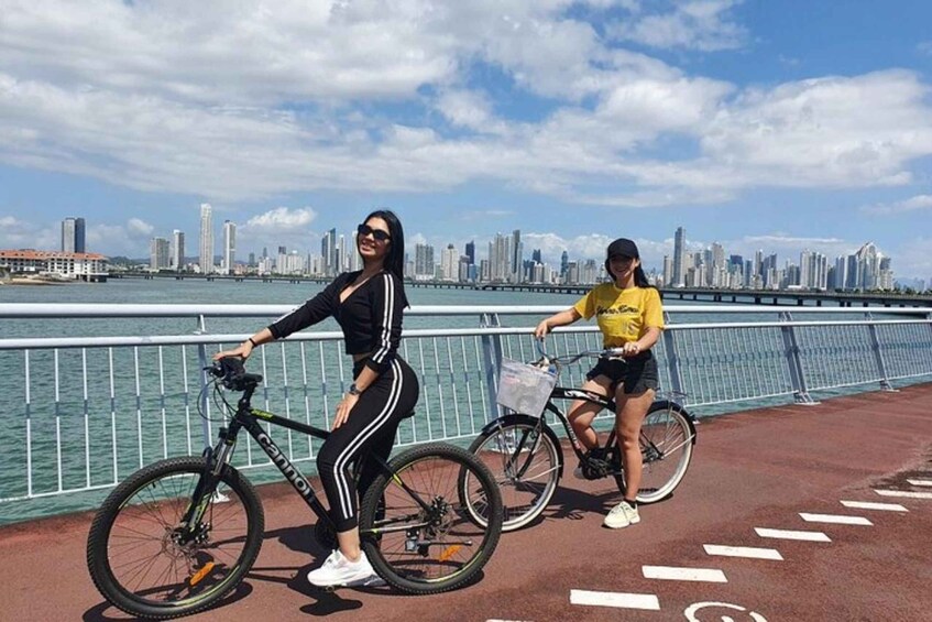 Two people on bicycles near a waterfront with a city skyline in the background. 2