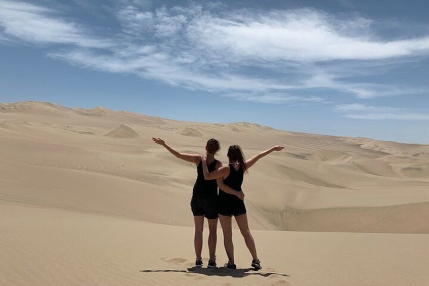 Two people standing on a sand dune with their arms outstretched, facing a vast desert landscape. 3