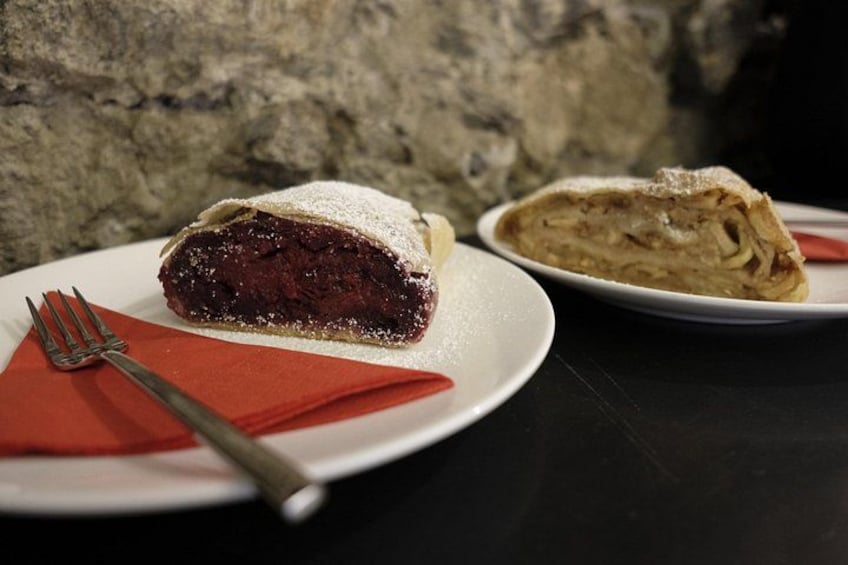 Una fetta di torta ciliegina su un piatto bianco con una forchetta, su un tovagliolo rosso, e un fagottino di pasta su un altro piatto. 2