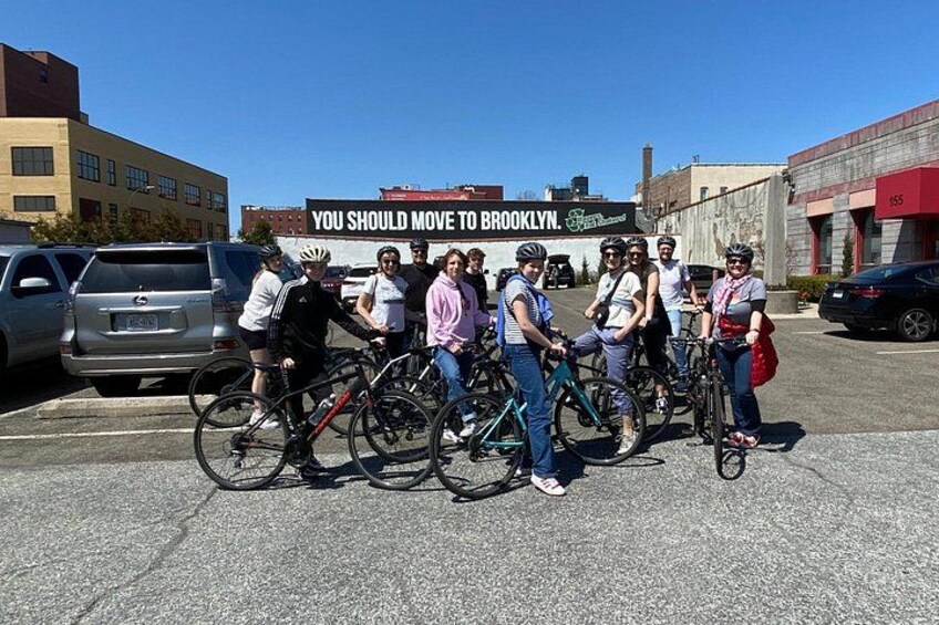 Een groep mensen met fietsen in een parkeerplaats, met een bord waarop staat 'VERHUIS NAAR BROOKLYN'. 4