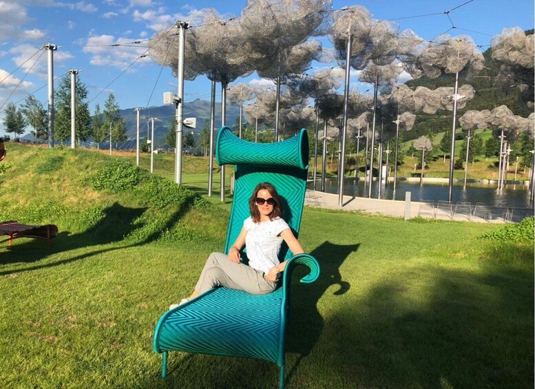 A person sitting on a green chair with a zigzag pattern, in a park with a lake and trees in the background. 5