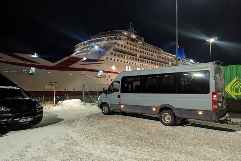 Een groot cruiseschip ligt aangemeerd in een haven, met een witte bestelwagen en een zwarte auto in de buurt. 3