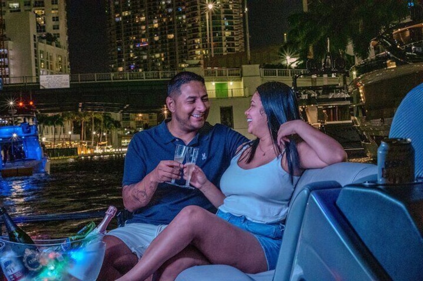 Dos personas en un bote por la noche, sosteniendo bebidas, con un horizonte urbano y agua de fondo. 5
