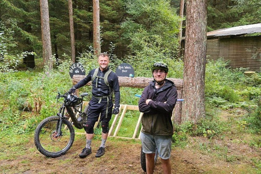 Two people standing next to a mountain bike in a forested area with a sign that reads 'Bike Park.' 5