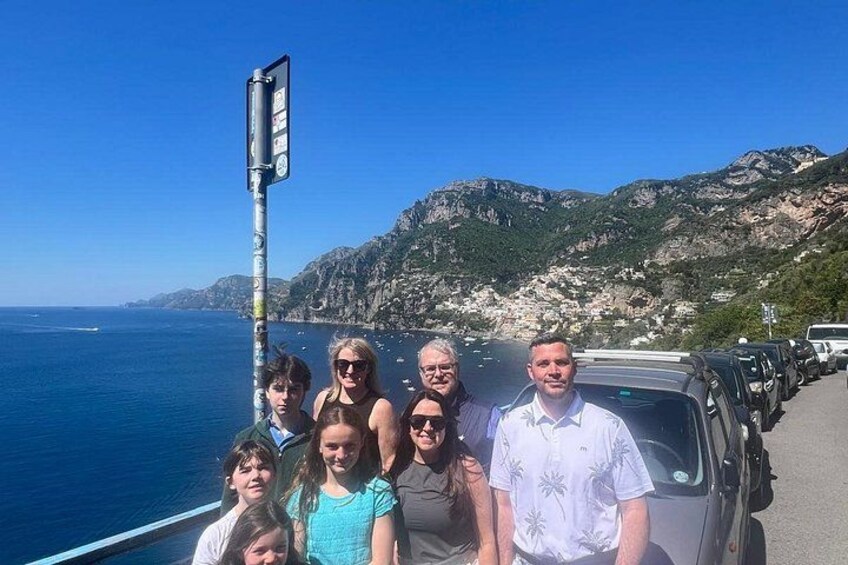 Eine Gruppe von Personen steht an einer Straße, im Hintergrund eine gebirgige Küstenlandschaft. 5