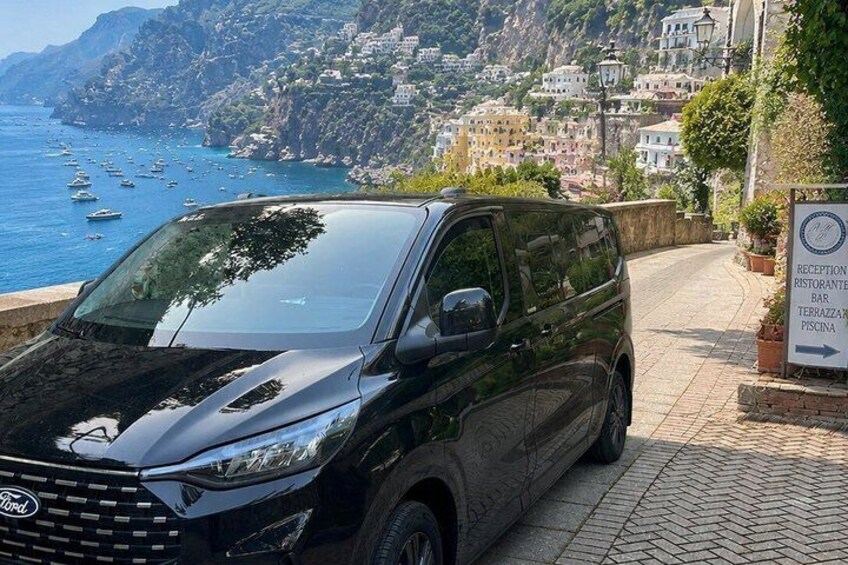 En sort Ford varevogn parkeret på en brostensbelagt sti med udsigt over kysten, et skilt og en bygning i baggrunden. 2
