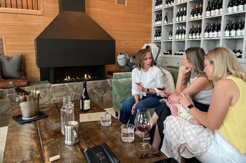 A group of people sitting around a wooden table with wine glasses and bottles, in front of a stone fireplace. 5