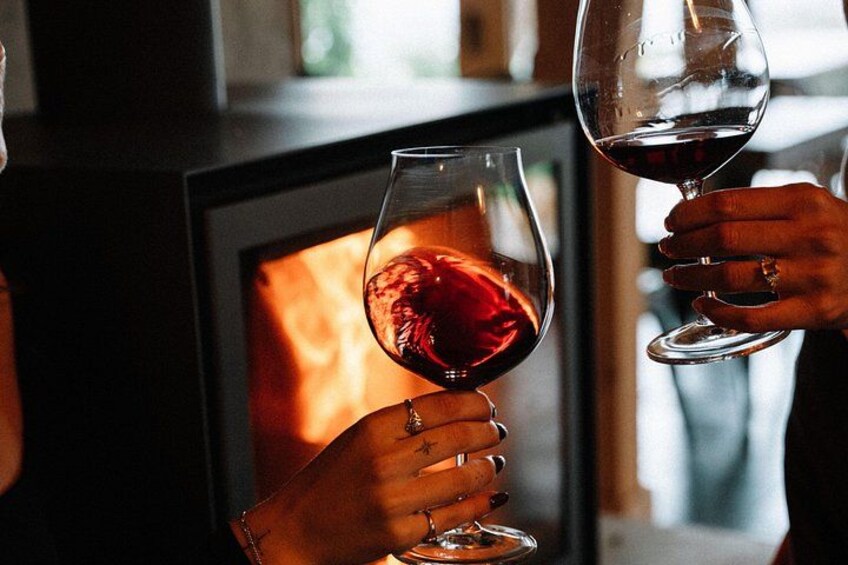 Two hands holding wine glasses with red liquid, in front of a fireplace. 3