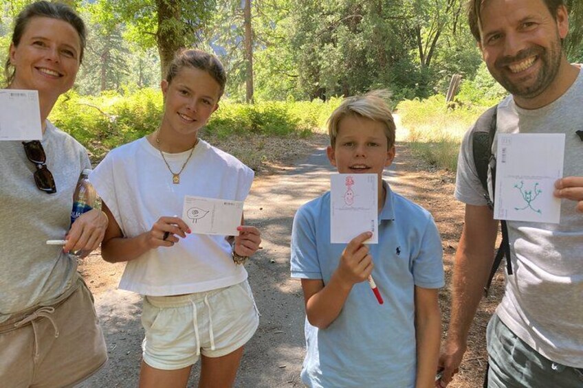 Four people holding papers with drawings, standing in a wooded area. 5