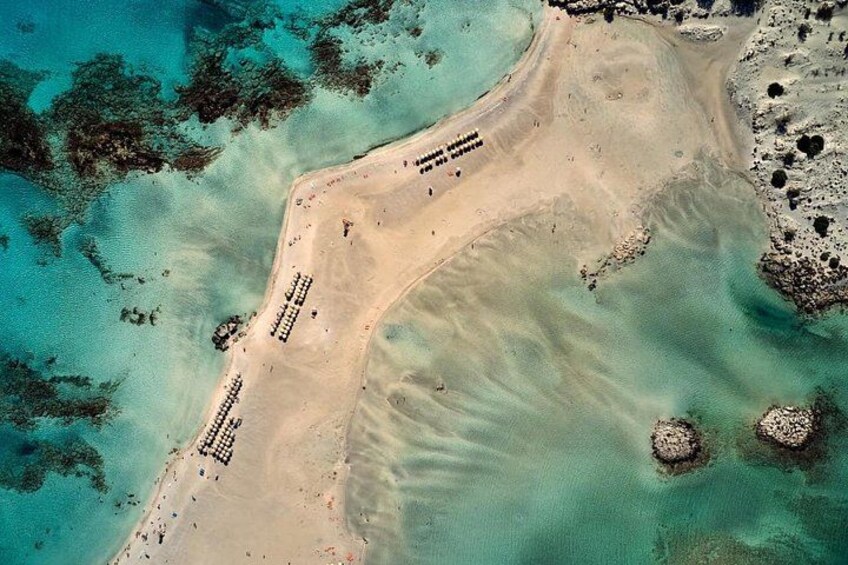Eine Vogelperspektive zeigt einen Strand mit klarem türkisblauem Wasser, sandigem Ufer und vereinzelten Felsen. 5