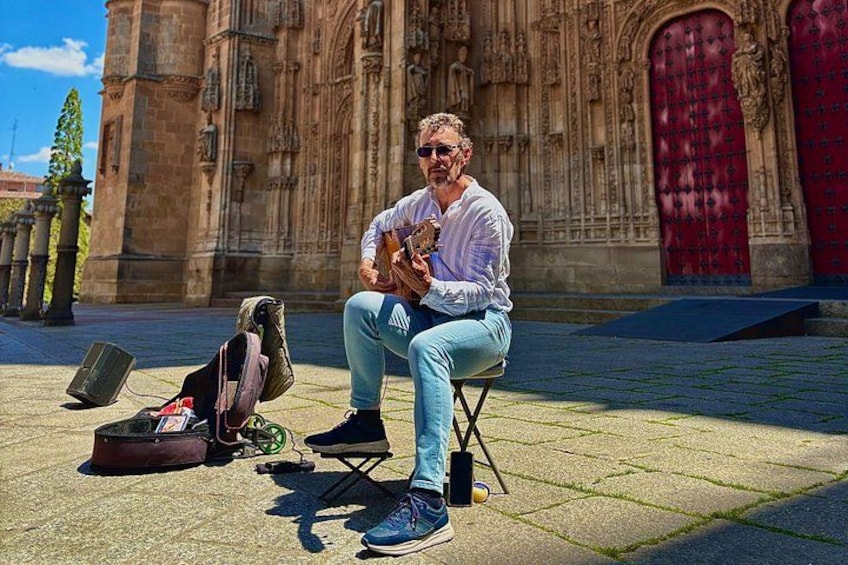 A person sitting on a stool playing a guitar in front of a historic building with intricate architecture. 3