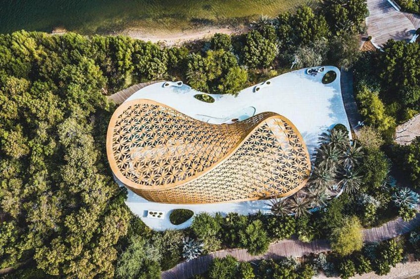 An aerial view of a unique architectural structure with a curved roof, surrounded by dense greenery and a beach. 4