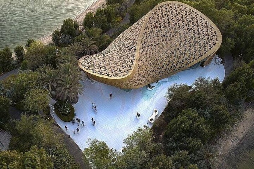A unique architectural structure with a patterned roof, surrounded by a landscaped area with trees and a body of water. 2