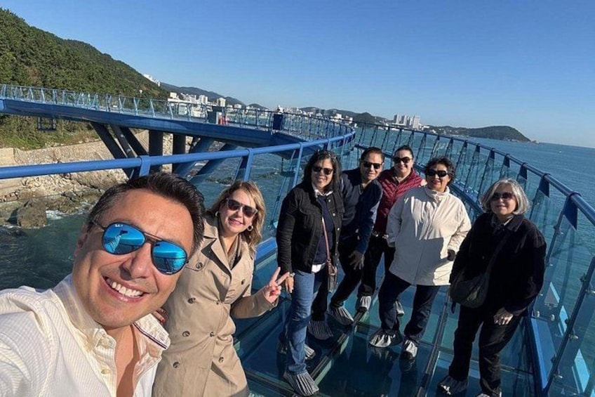 A group of people on a glass bridge over water with a coastal landscape in the background. 3