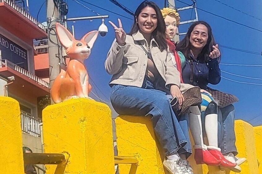 Three people sitting on a yellow fence with a statue of a fox in the background. 3