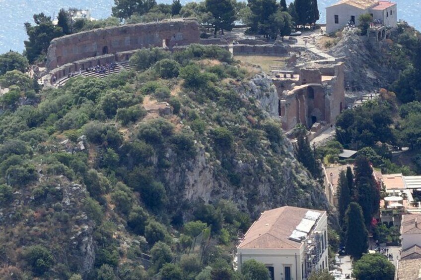 Un sitio histórico con ruinas, que incluye un teatro y edificios antiguos, rodeado de vegetación y con vistas a un cuerpo de agua. 3