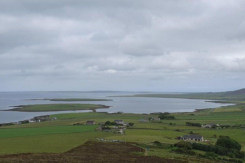 Un paysage côtier avec des champs, des maisons et une étendue d’eau, le tout sous un ciel nuageux. 2