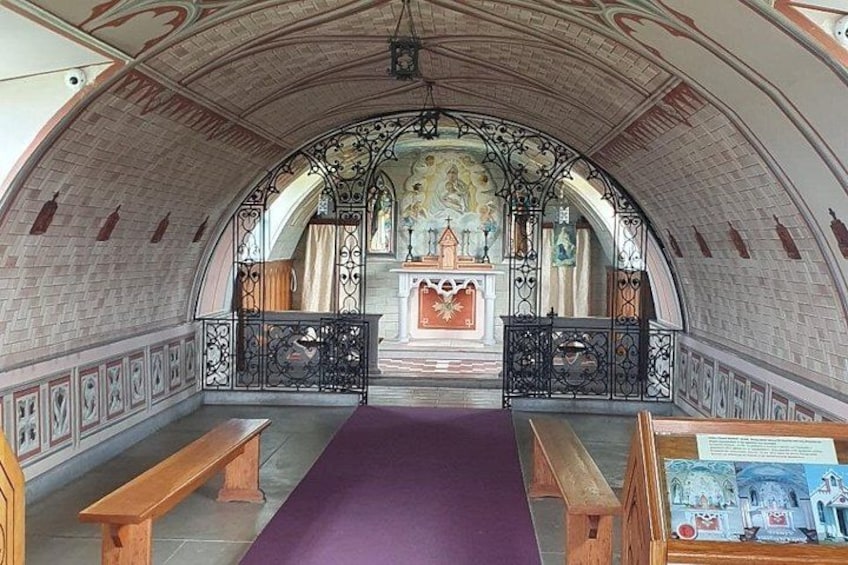 Ein Innenraum mit Bogen, einem violetten Teppich, Bänken und einem zentralen Altar. 2