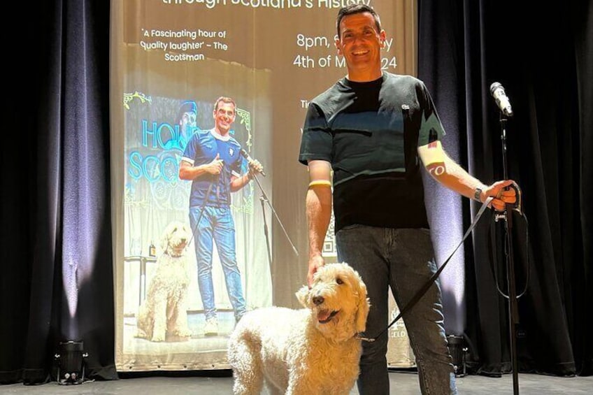 A person on stage with a microphone, a dog, and a backdrop with text. 5