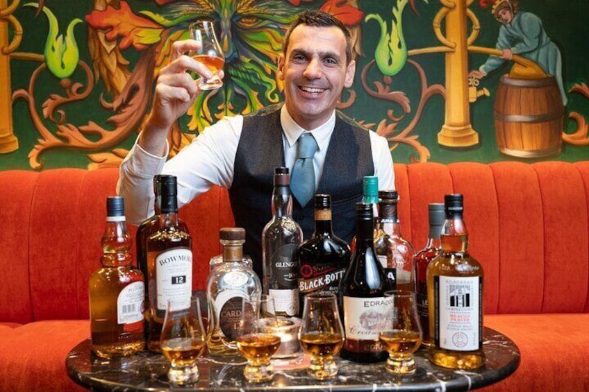 A person holding a glass of whiskey with various bottles of whiskey and a bottle of gin on a table. 1