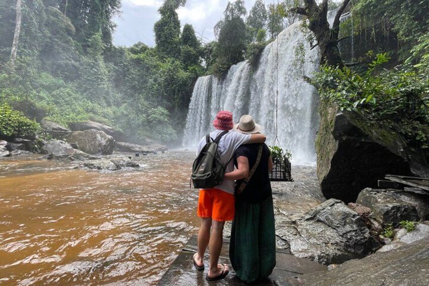 A waterfall with a couple standing on a rocky ledge. 1