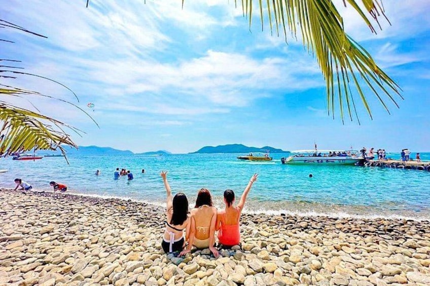 A beach with people sitting on pebbles, a clear blue sky, and boats in the water. 1
