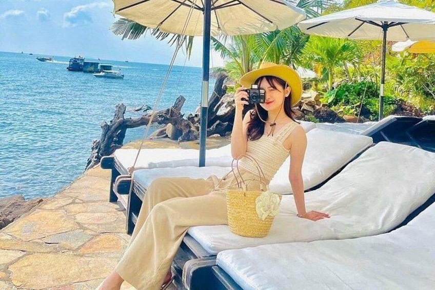 A person sitting on a lounge chair by the water, holding a camera. 5