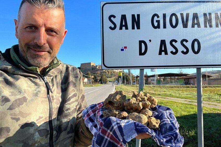 A person holding a pile of rocks with a sign that reads 'SAN GIOVANNI D'ASSO' in the background. 3