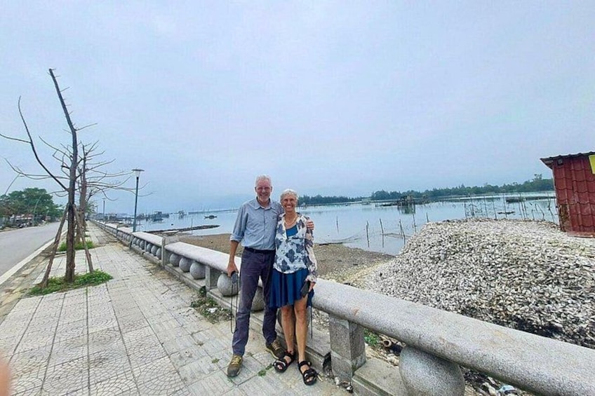 A man and woman standing on a concrete walkway by a body of water, with a pile of gravel and a small red structure in the background. 5