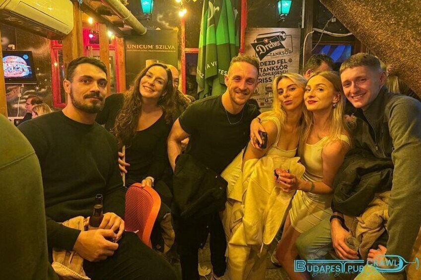 A group of people in a dimly lit indoor setting, possibly a bar or club, with a sign that reads "UNICUM SZILVA" and "Budapest Pub Crawl" visible in the background. 5
