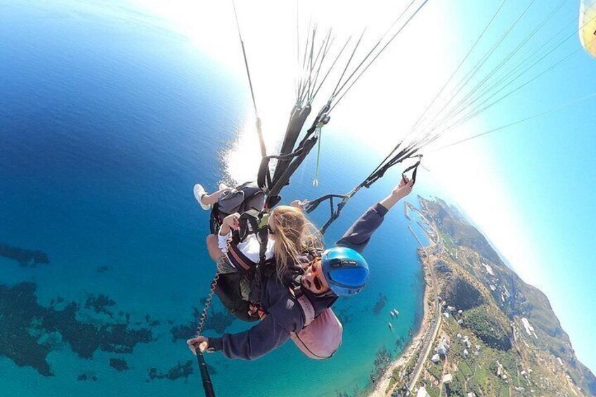 Eine Person gleitet mit einem Paragleiter über eine Küstenlandschaft mit klarem, blauem Wasser und hügeligem Gelände darunter. 2
