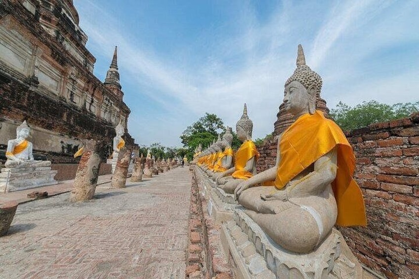 Filas de estatuas de Buda sentadas, con túnicas anaranjadas, frente a un muro de ladrillo y antiguas estructuras de templo. 3