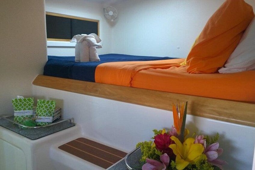 En køjeseng med orange sengetøj, et lille vindue, et natbord med en vase blomster og en reol med genstande. 4