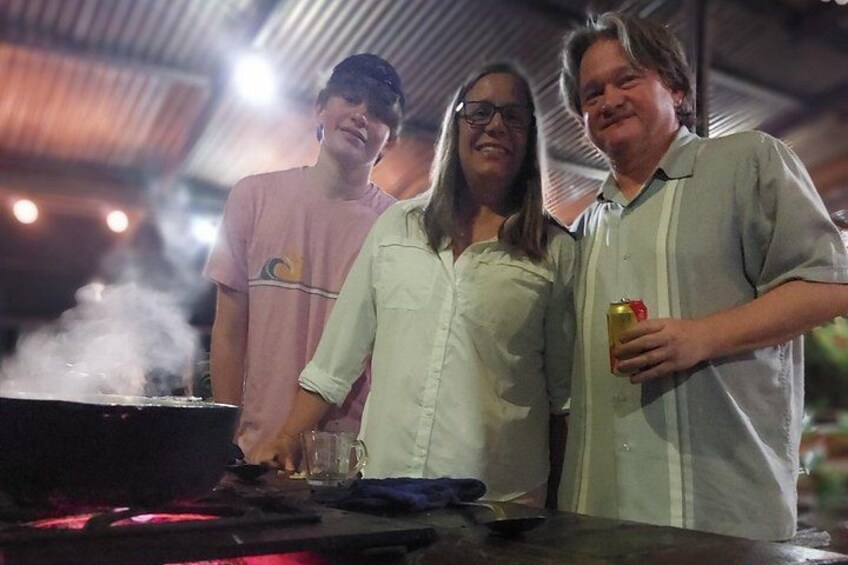 Trois personnes se tiennent près d’un barbecue où de la fumée s’échappe, une table avec un verre et une bouteille. 2