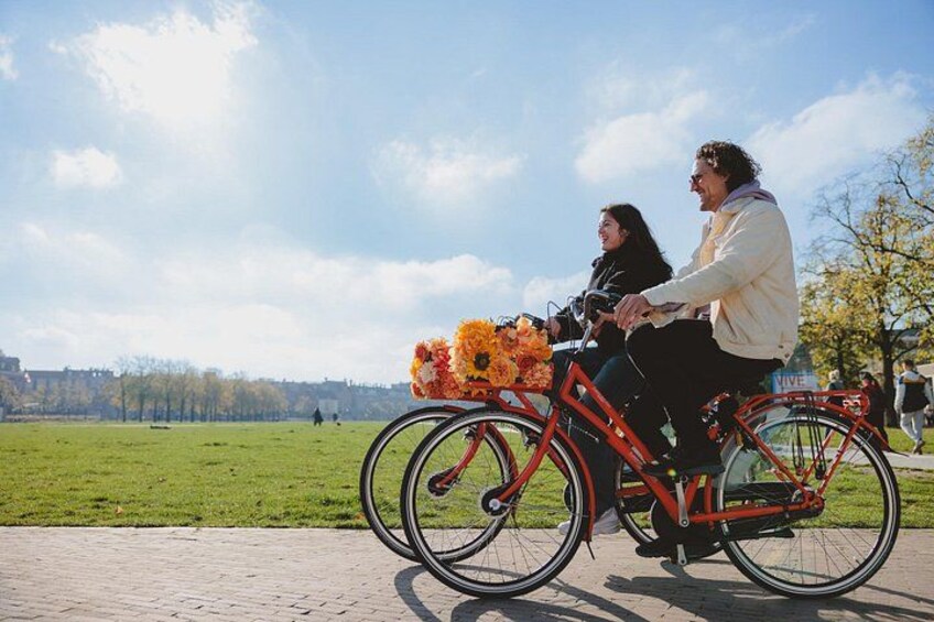 Two people riding a bicycle with a basket of flowers, on a paved path with a grassy field and trees in the background. 4